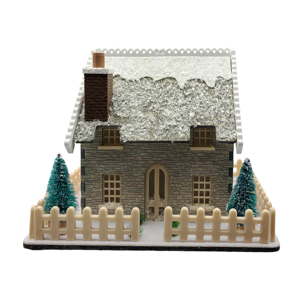 A miniature winter cottage model with a snow-covered roof, stone-textured walls, a small chimney, a beige picket fence, and two small evergreen trees, shown on a plain white background.