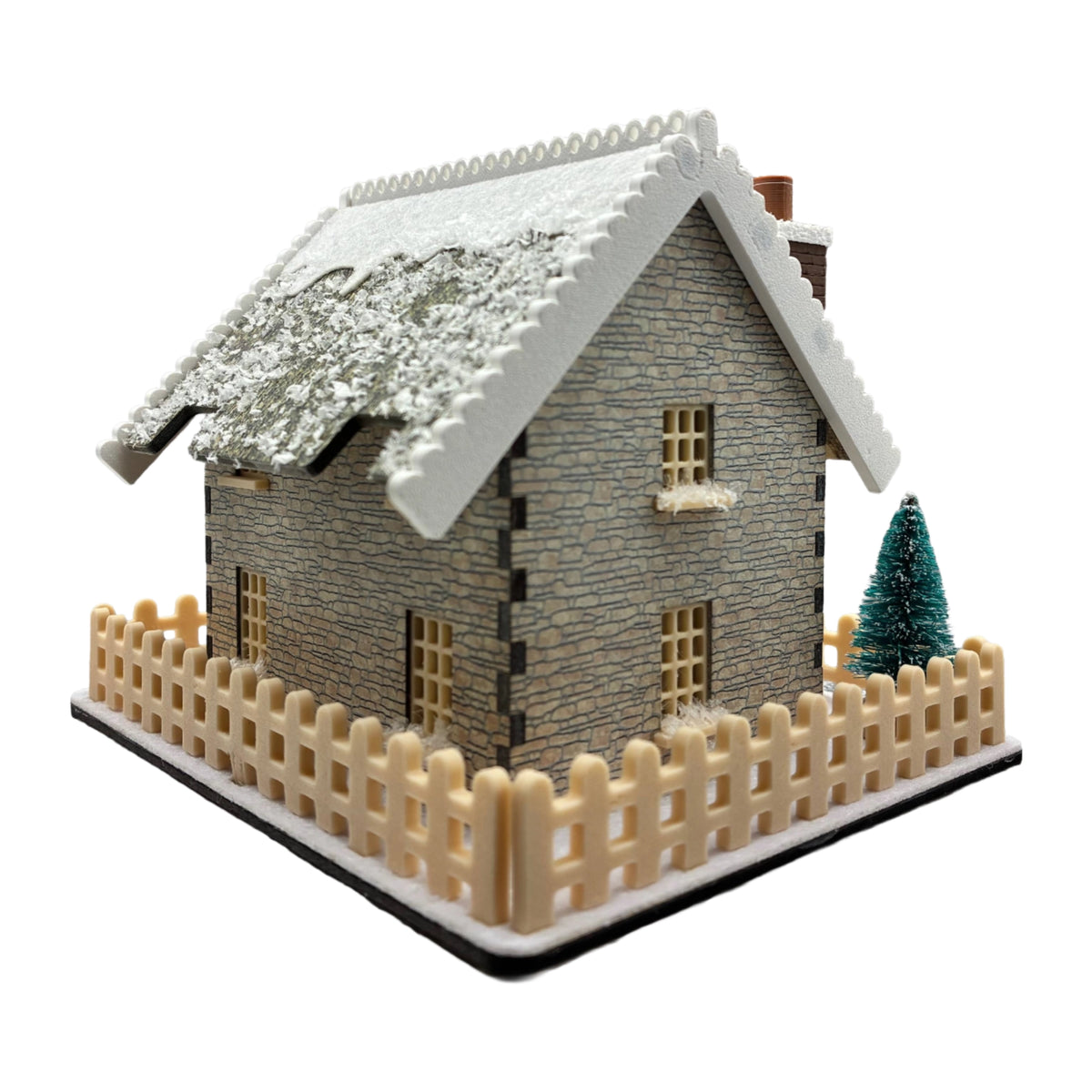 A miniature winter stone cottage model with a snowy roof, beige picket fence, and a small green tree, shown on a plain white background.