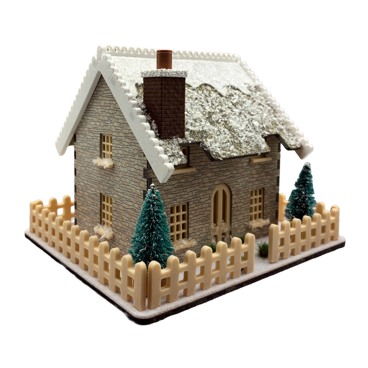 A miniature winter cottage model with a snow-covered roof, stone-textured walls, a small chimney, a beige picket fence, and two small evergreen trees, shown on a plain white background.